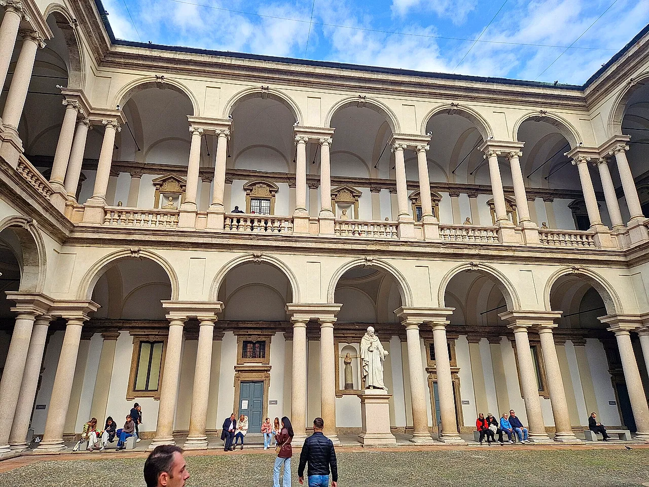 Der Hof des Palazzo Brera mit weißen Arkaden auf zwei Geschossen und einer weißen Statue auf einem Sockel in der Mitte; im Vordergrund stehen Besucher