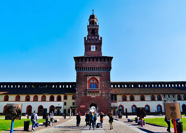 La torre d'ingresso del Castello Sforzesco vista dal cortile, con il portale ad arco e gli edifici color crema con finestre ad arco rosse ai lati, e visitatori che camminano sul selciato.