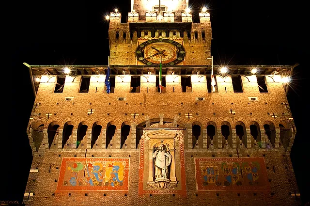Ein nachts beleuchteter Backsteinturm des Castello Sforzesco mit einer Uhr im oberen Teil und einer Steinstatue in einer mittleren Nische zwischen zwei verzierten Feldern vor dunklem Himmel