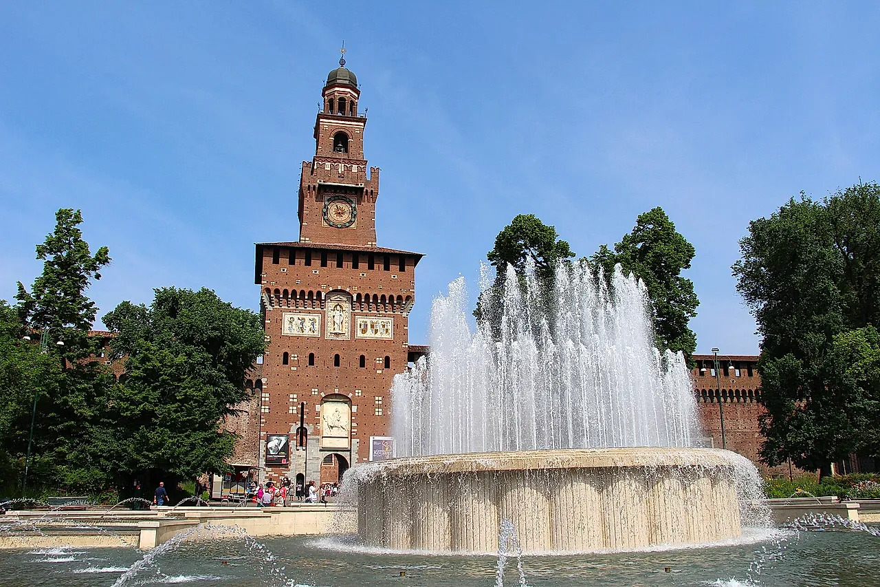 La torre del reloj del Castillo Sforzesco vista al otro lado de una fuente, con los chorros de agua elevándose delante de los muros de ladrillo rojo bajo un cielo azul despejado.
