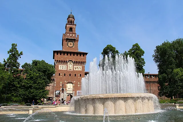 Der Uhrturm des Castello Sforzesco über einen Brunnen hinweg gesehen, dessen Wasserstrahlen vor den roten Backsteinmauern unter klarem blauem Himmel aufsteigen