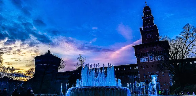 Das Castello Sforzesco in der Dämmerung, sein Backsteinturm und die Mauern als Silhouette vor blauem und rosafarbenem Himmel, mit einem beleuchteten Brunnen im Vordergrund