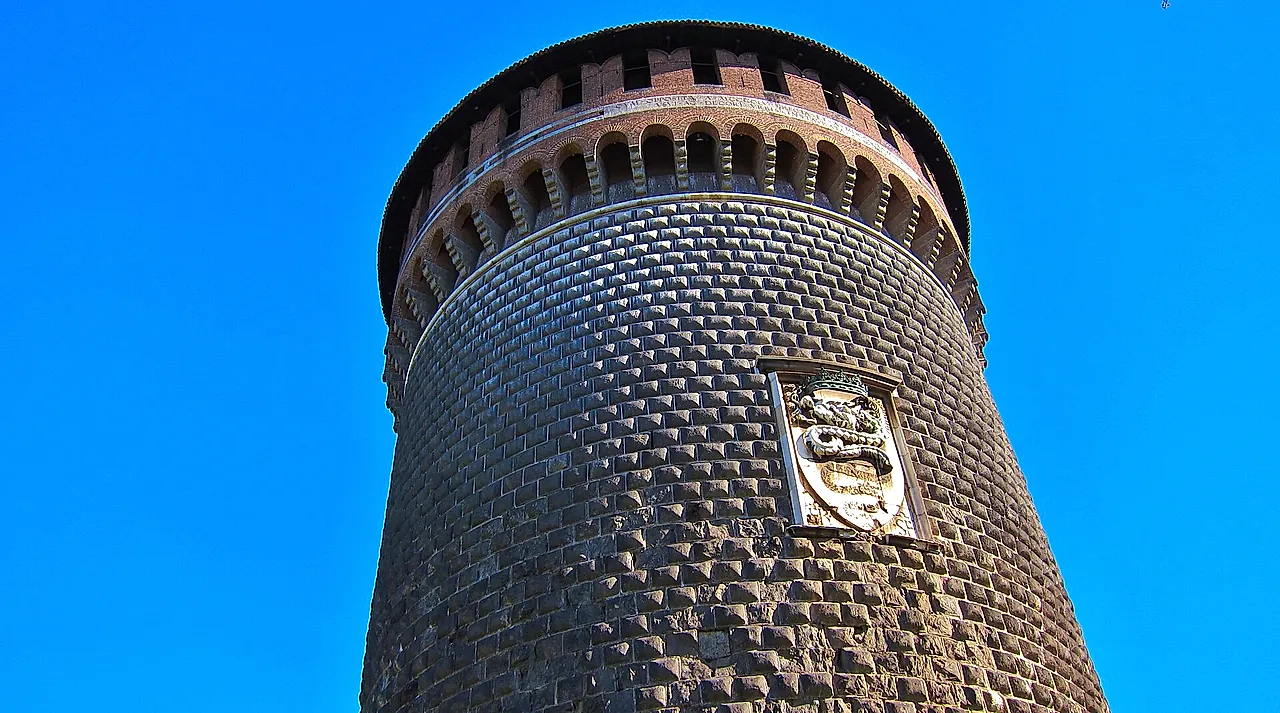 La torre redonda del Castillo Sforzesco, construida con piedra oscura y ladrillo, vista desde abajo bajo un cielo azul despejado, con un escudo de armas de piedra tallada en el muro.