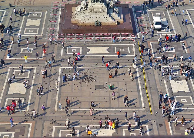 Blick von oben auf die Piazza del Duomo vom Dach der Kathedrale: das helle geometrische Pflaster, eine Menge von Besuchern und Tauben und am oberen Bildrand ein steinernes Denkmal