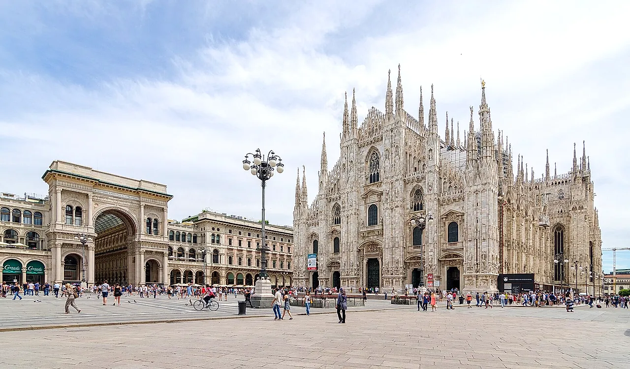 El arco de la Galleria Vittorio Emanuele II a la izquierda y la fachada de la catedral a la derecha, al otro lado de una amplia plaza, con peatones caminando por la plaza.
