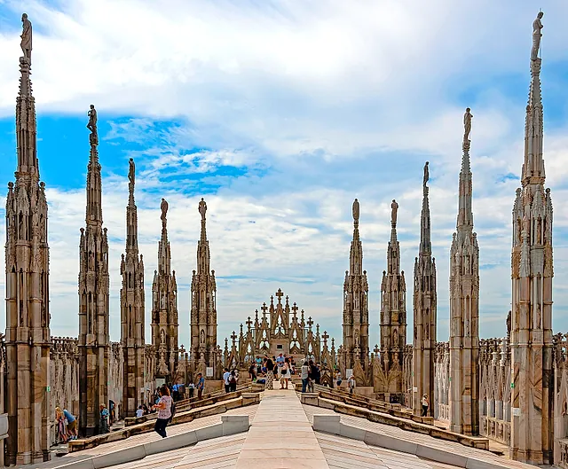 Die Dachterrassen des Doms, gesäumt von Reihen marmorner Türmchen mit Statuen auf der Spitze, mit einem Mittelweg zur Fassade unter bewölktem Himmel und einigen Besuchern auf dem Pfad