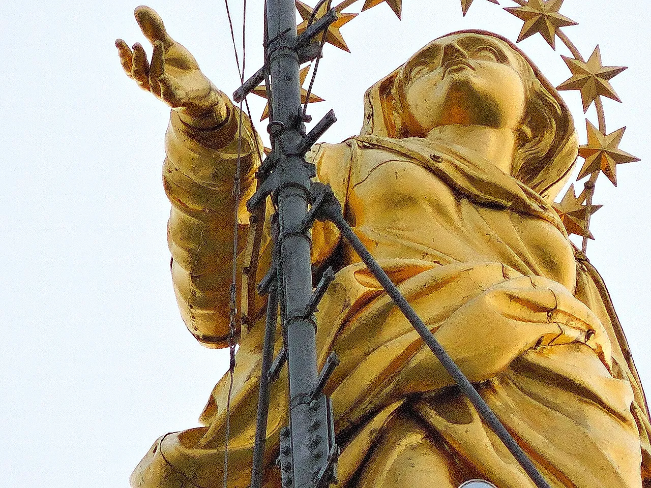La estatua dorada de la Madonnina sobre la aguja de la catedral, vista desde abajo contra un cielo pálido, con el brazo extendido y la corona de estrellas, y un pararrayos metálico con cables delante.