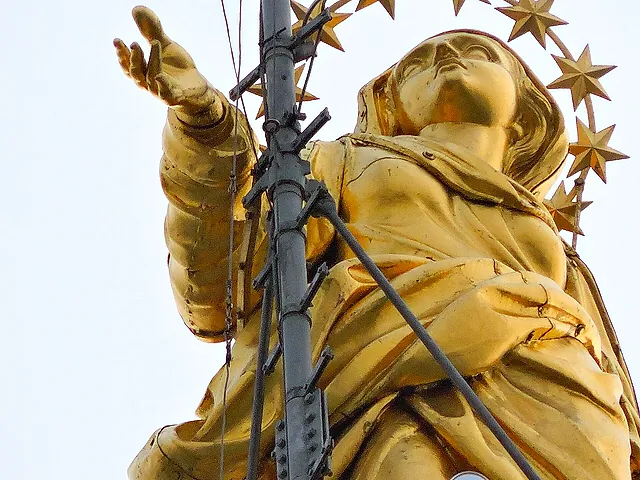 Die vergoldete Statue der Madonnina auf der Spitze des Domtürmchens, von unten vor blassem Himmel gesehen, mit ausgestrecktem Arm und Sternenkranz und einem Blitzableiter mit Kabeln davor