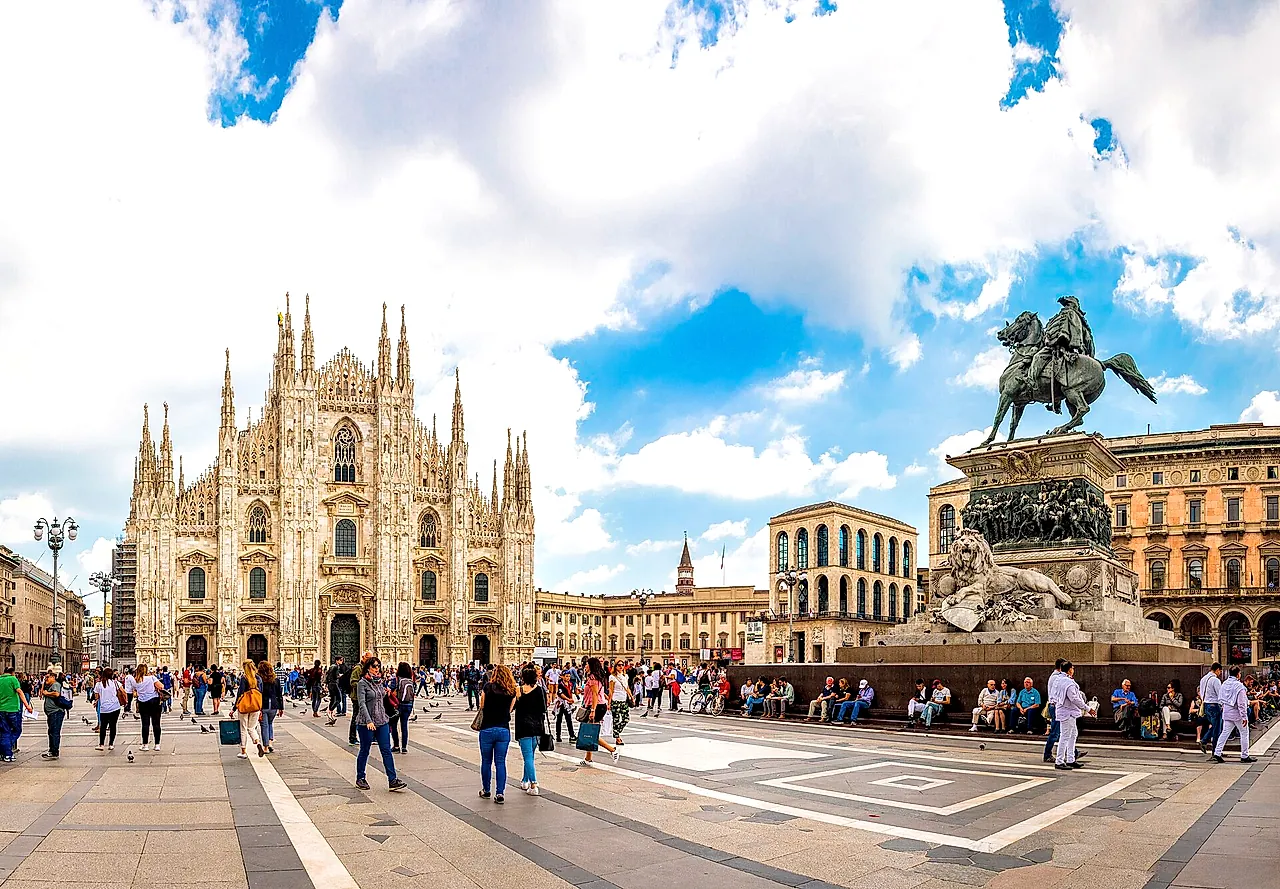 Die Piazza del Duomo mit der Kathedrale links und dem Reiterstandbild Viktor Emanuels II. rechts, weitem Himmel mit Wolken und Passanten im Vordergrund