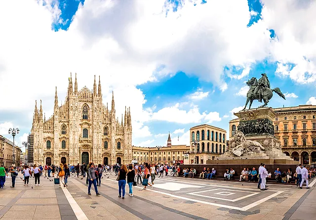 Piazza del Duomo con la cattedrale a sinistra e la statua equestre di Vittorio Emanuele II a destra, un ampio cielo con nuvole e passanti in primo piano.