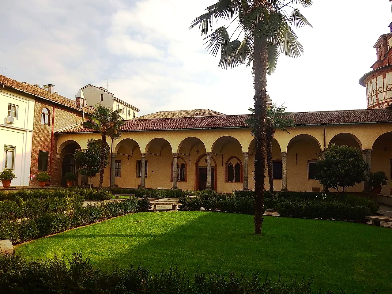 Der Kreuzgang von Santa Maria delle Grazie: eine cremefarbene Arkade um einen Rasen, eine hohe Palme in der Mitte und geschnittene Hecken entlang der Beete