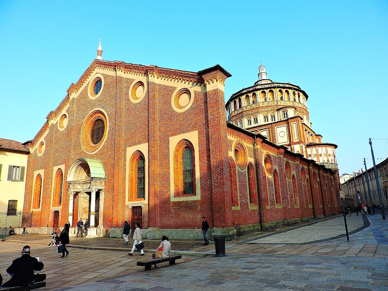 Die Backsteinfassade von Santa Maria delle Grazie an einem sonnigen Platz, mit einem hellen Steinportal, Rundbogenfenstern und dem runden Kuppeltambour rechts