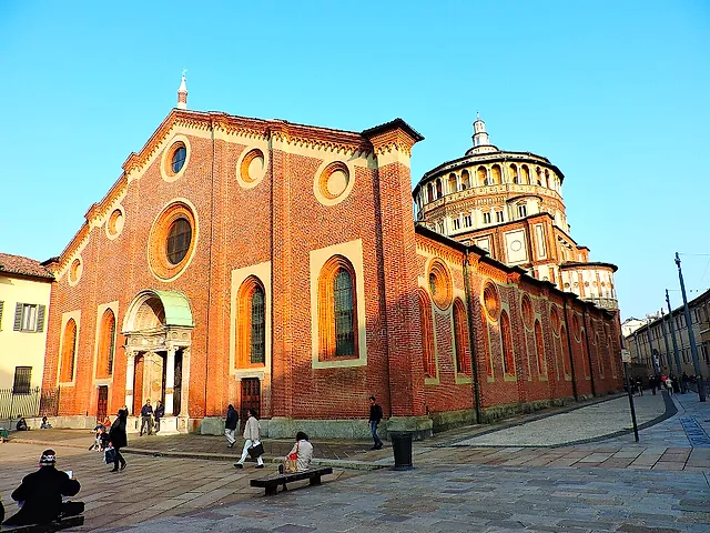 Die Backsteinfassade von Santa Maria delle Grazie an einem sonnigen Platz, mit einem hellen Steinportal, Rundbogenfenstern und dem runden Kuppeltambour rechts