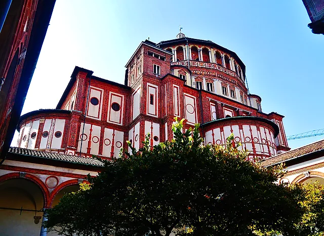 Die rote Backsteinapsis von Santa Maria delle Grazie von Norden gesehen, mit einem Kuppeltambour über Reihen runder Fenster, eingerahmt von einem belaubten Baum im Vordergrund