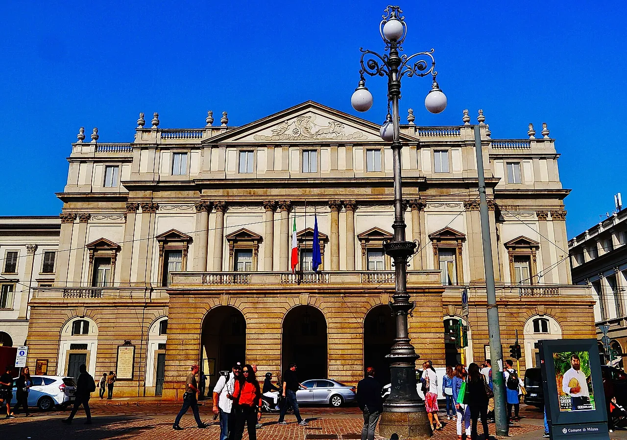 La fachada neoclásica del teatro de la ópera de La Scala, con columnas, un frontón triangular y entradas en arco a nivel de la calle, vista desde la plaza con transeúntes.