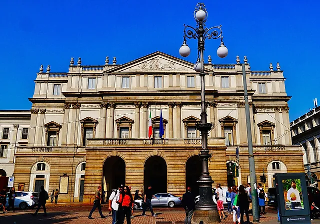 Die klassizistische Fassade des Opernhauses La Scala mit Säulen, einem Dreiecksgiebel und Bogeneingängen auf Straßenebene, vom Platz aus gesehen, mit Passanten