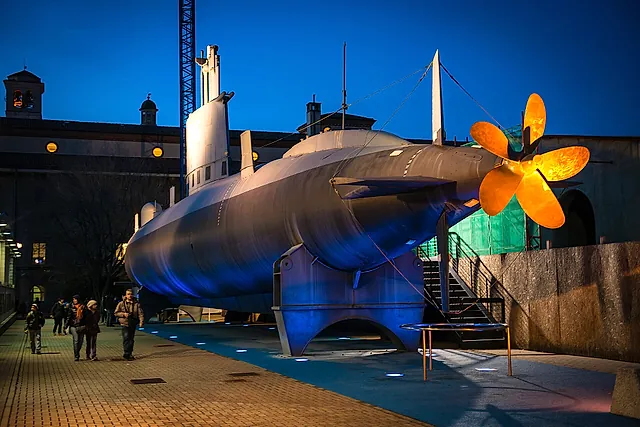 Das U-Boot Enrico Toti in der Dämmerung im Freien ausgestellt, sein grauer Rumpf von blauen Scheinwerfern angestrahlt und sein goldener Propeller rechts leuchtend, mit einigen Besuchern links